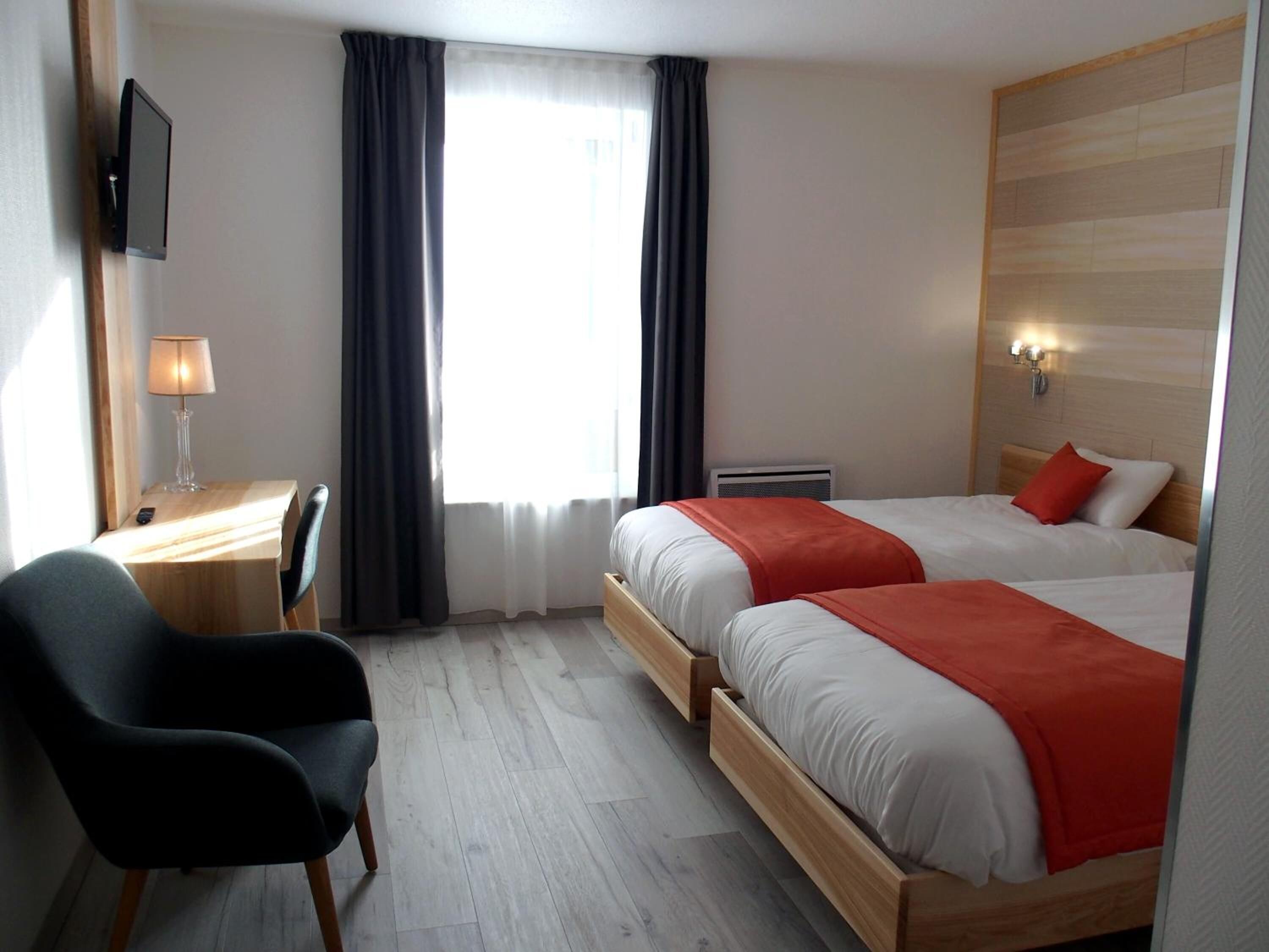 Ein Hotelzimmer mit zwei Betten, einem Schreibtisch mit Lampe, einem Sessel und einem an der Wand montierten Fernseher.