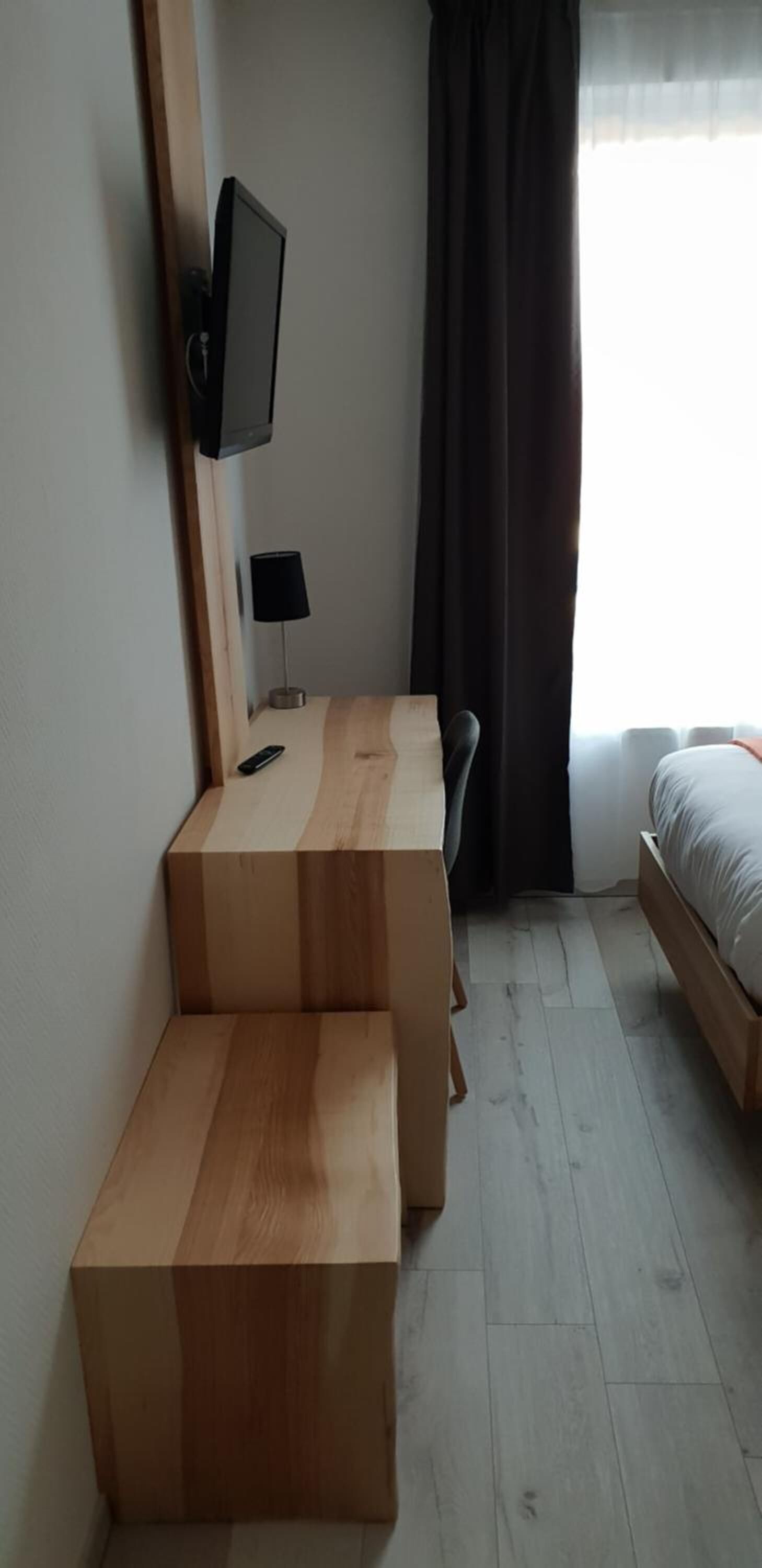 Een kamer met een bed, een aan de muur bevestigde televisie, een bureau met een lamp en een raam met gordijnen.
