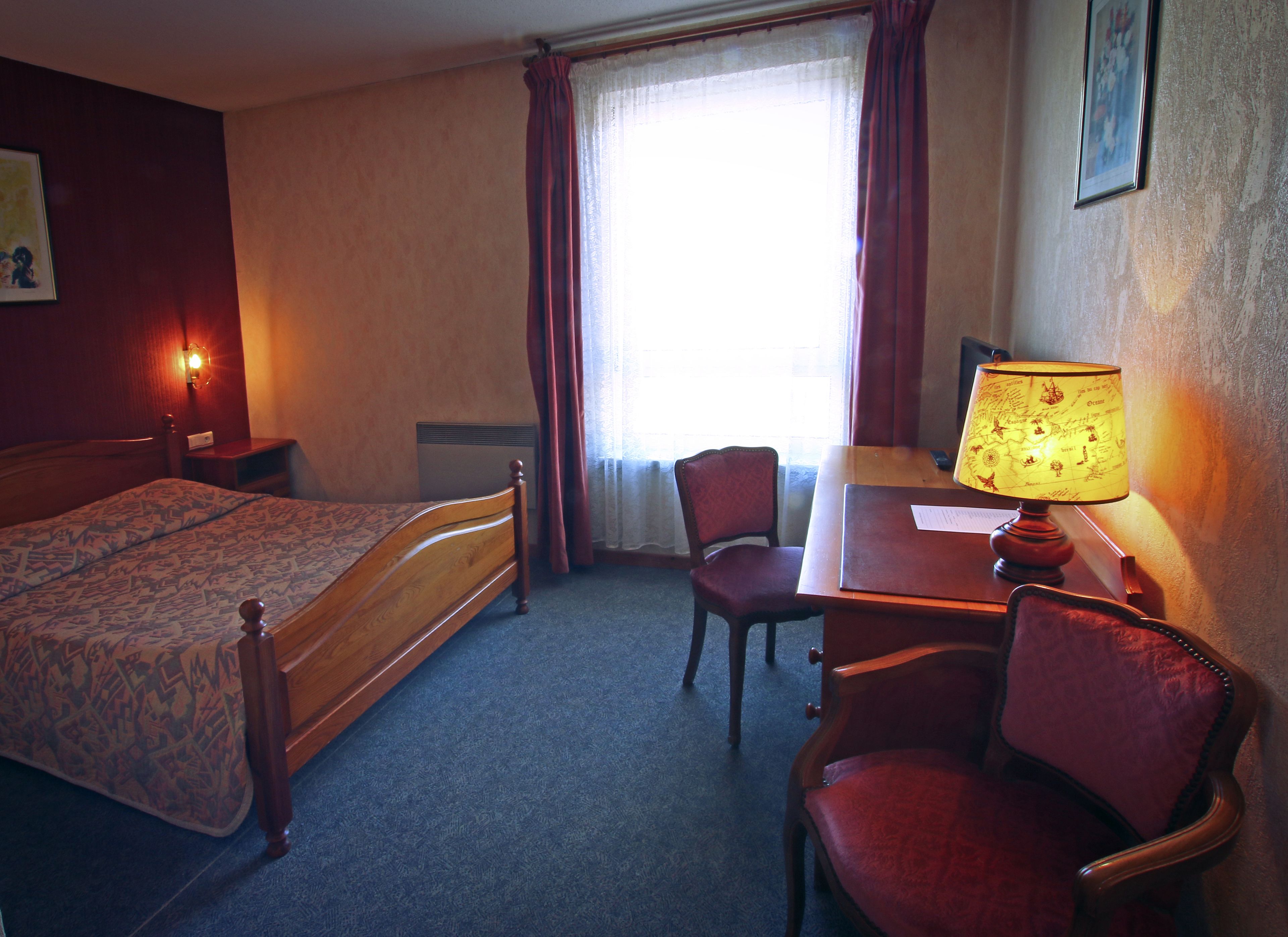 Une chambre d’hôtel avec un lit, un bureau, une chaise, une lampe, une table de chevet et une fenêtre avec des rideaux.