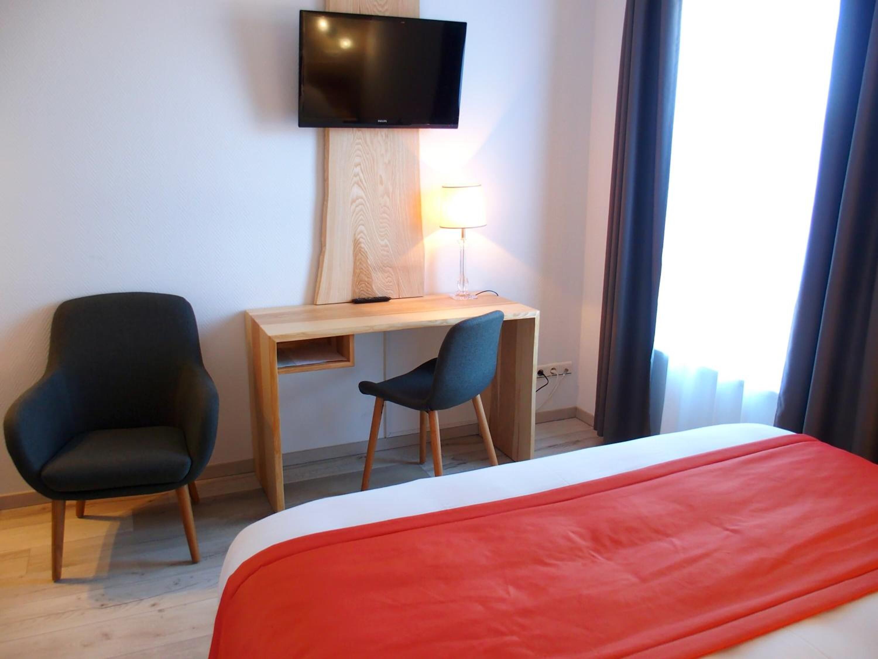 Une chambre d’hôtel avec un lit, un bureau, deux chaises, une télévision et une fenêtre avec des rideaux.