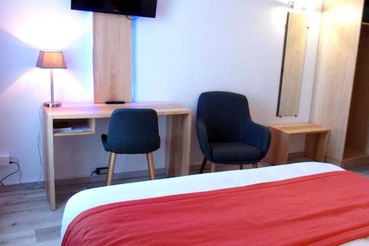 A hotel room with a bed, two chairs, a desk, a TV, and a wardrobe.