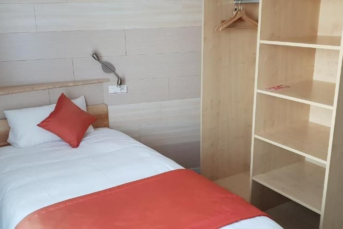 A bedroom with a wooden headboard, a bed with white bedding and a red pillow, a bedside table with a lamp, and a wooden wardrobe with shelves.