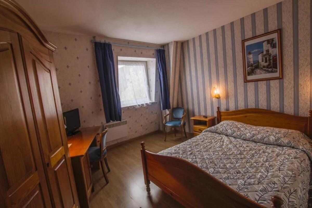 Ett sovrum med en säng i trä, ett skrivbord med en dator, en stol, ett fönster med gardiner och en vägglampa.