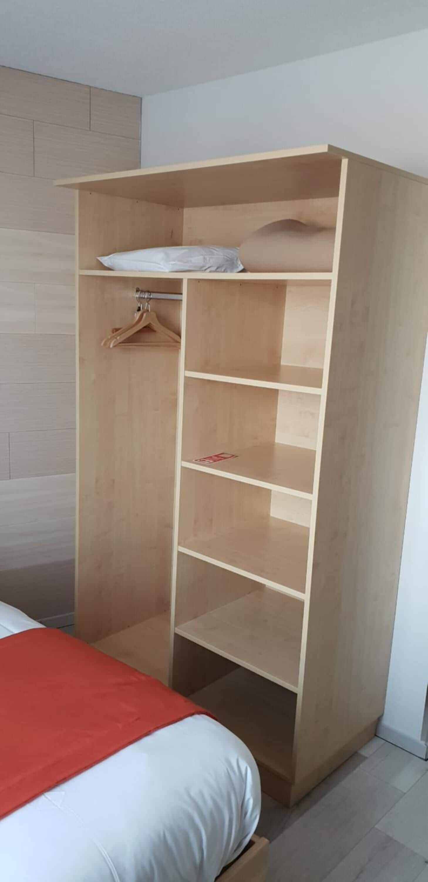 A wooden wardrobe with multiple shelves and a hanging rod, located next to a bed with a red blanket.
