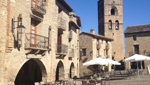 Una plaza adoquinada con edificios de piedra, una torre del reloj y mesas al aire libre con sombrillas.