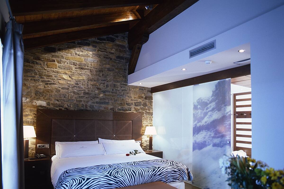 A bedroom with a stone wall, a wooden headboard, a zebra-patterned bedspread, a bench, and a nightstand with a lamp.