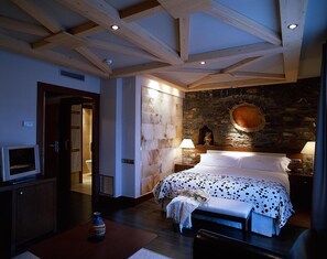 Um quarto com parede de pedra, cama com colcha de bolinhas, teto de madeira e uma televisão sobre um móvel.