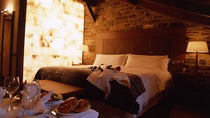 Un cuarto acogedor con pared de piedra, techo de madera, cama con sábanas blancas, mesita de noche con lámpara y una mesa para el desayuno con croissants y café.