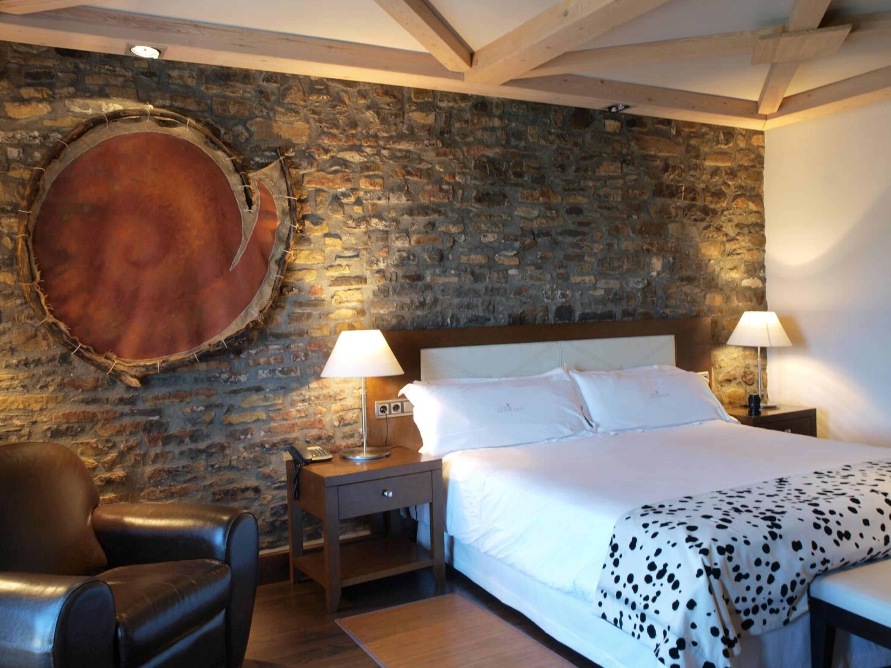 A bedroom with a stone wall, a round wooden object, a bed with white bedding, two bedside lamps, a wooden nightstand, a leather chair, and a wooden table.