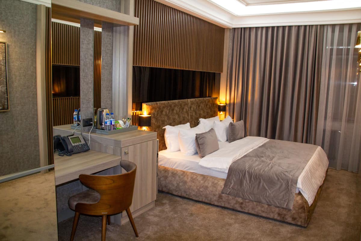 A hotel room with a bed, bedside tables, a desk, a chair, and a large window with curtains.