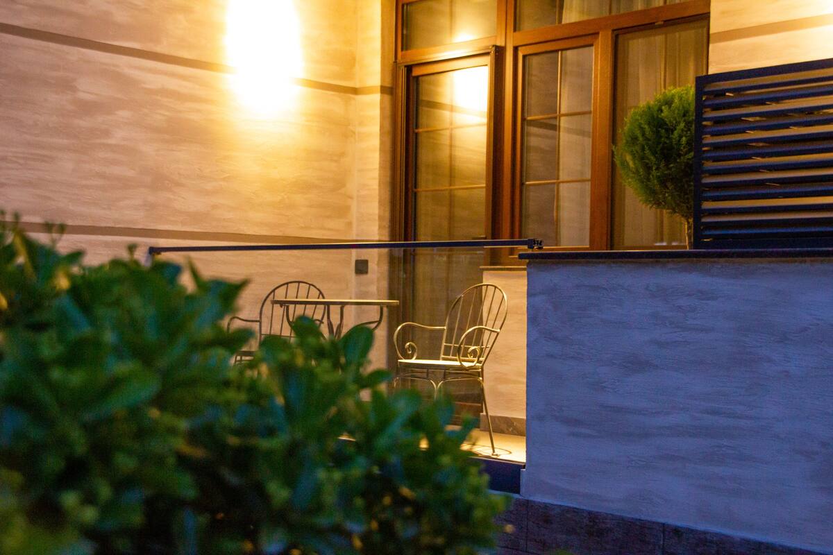 A balcony with metal chairs and a small potted plant, a wooden railing, and a window with curtains.