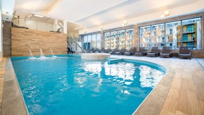 Une piscine intérieure dotée d’une terrasse en bois, de chaises longues et offrant une vue sur l’extérieur du bâtiment grâce à de grandes fenêtres.