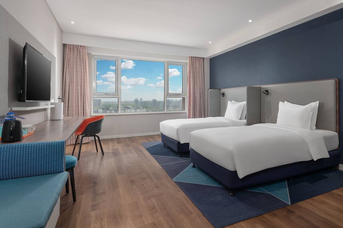 Habitación de hotel con dos camas, televisión, escritorio con silla y vista a la ciudad.