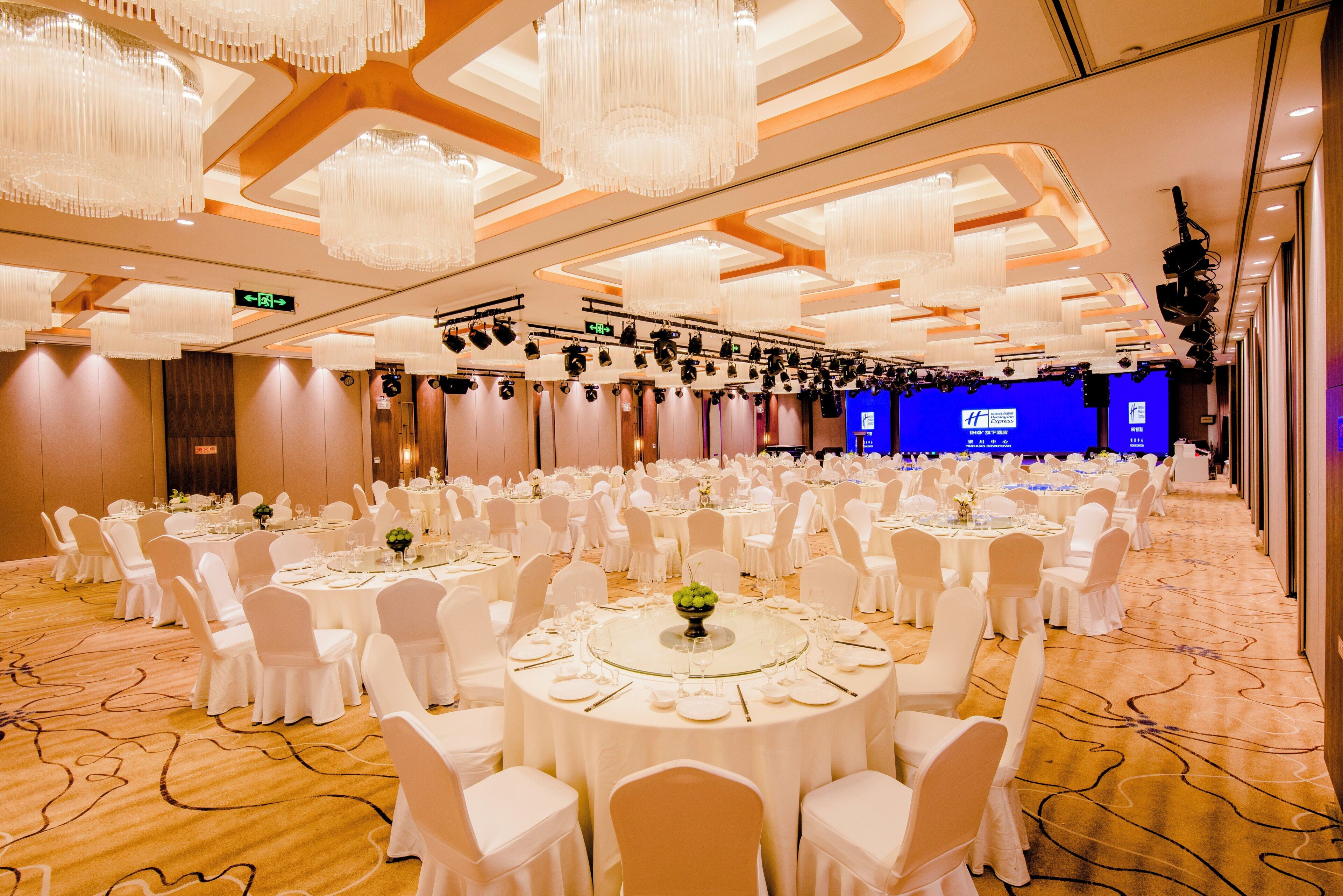 A banquet hall with round tables, white chairs, and chandeliers.