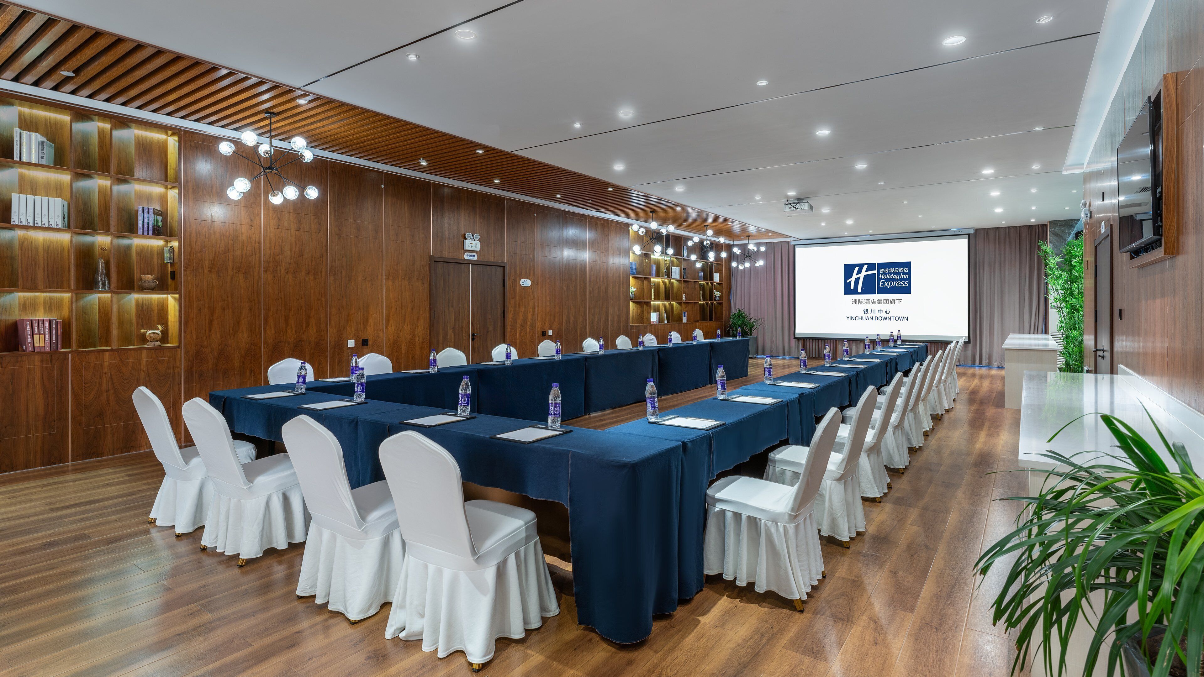 A conference room with long tables, white chairs, and a projector screen displaying a logo.