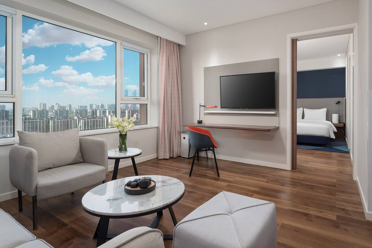 Una habitación de hotel moderna con vista a la ciudad, un televisor de pantalla plana, una cama y una pequeña zona de estar con una mesa y sillas.