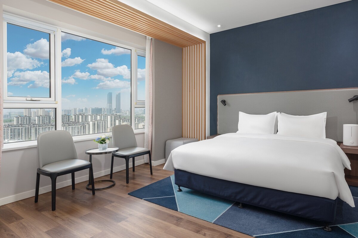 Une chambre d’hôtel avec un grand lit, un bureau avec une chaise, une fenêtre donnant sur la ville et une table de chevet avec une lampe.