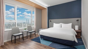Habitación de hotel con una cama grande, un escritorio con una silla, una ventana con vista a la ciudad y una mesita de noche con una lámpara.
