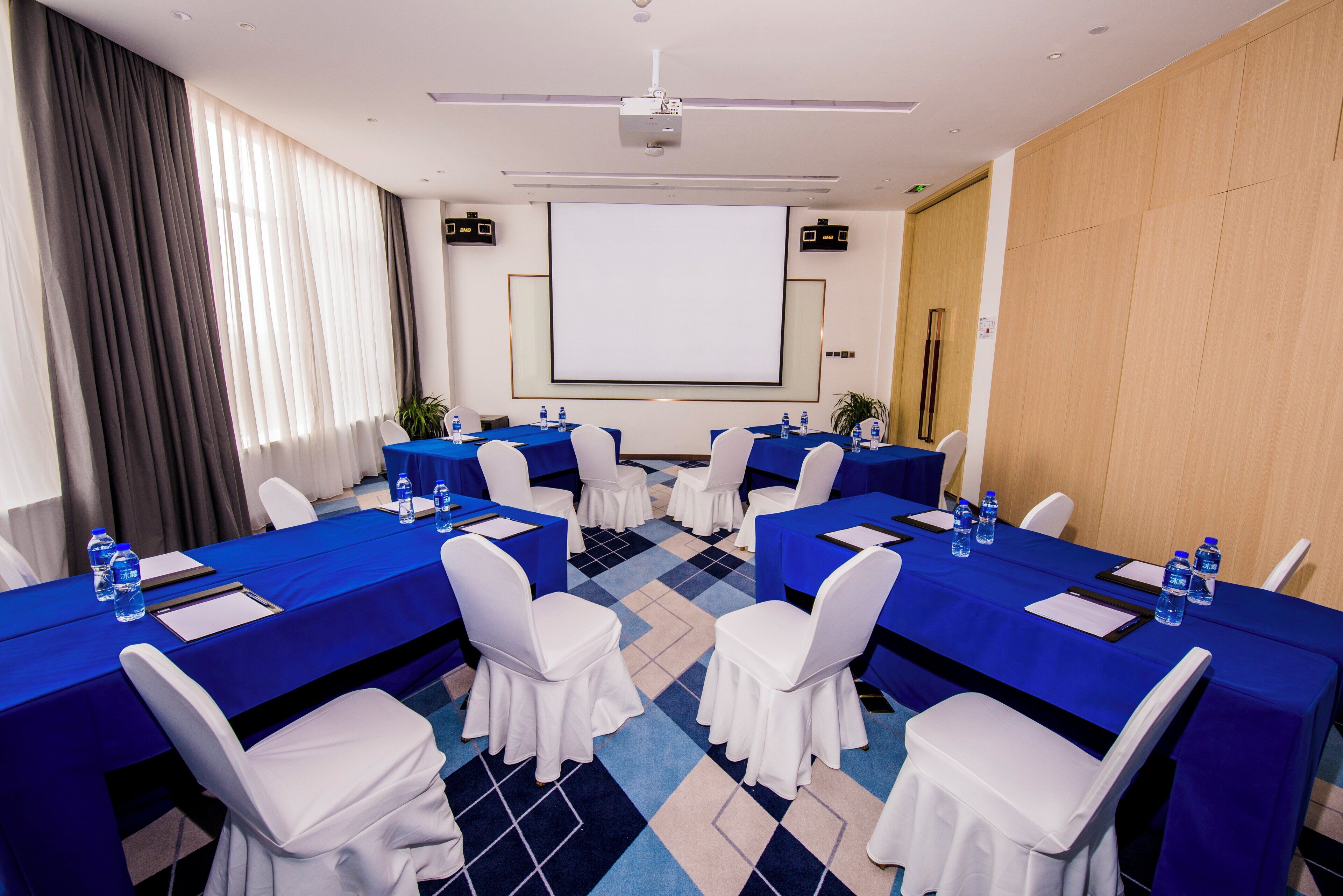 A conference room with a large screen, multiple tables with chairs, and water bottles on the tables.
