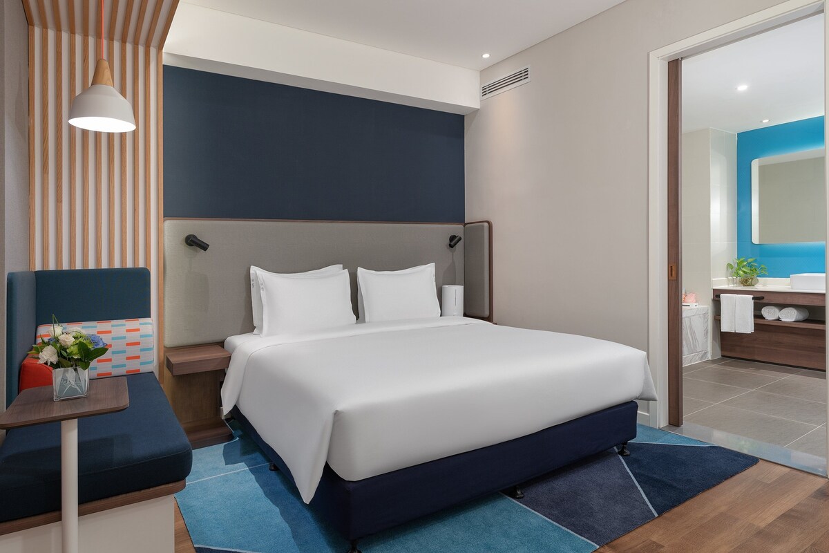 Une chambre d’hôtel moderne avec un grand lit, une table de chevet avec une lampe, un petit coin salon et une salle de bain visible à travers une porte ouverte.