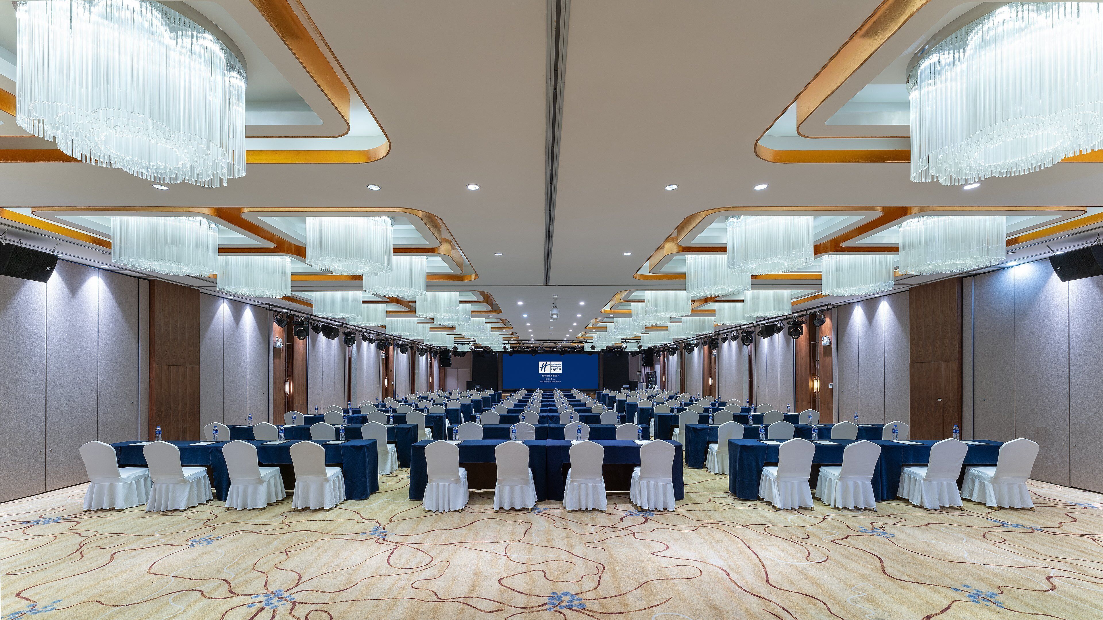 A conference room with rows of tables and chairs, a stage with a blue backdrop, and chandeliers.