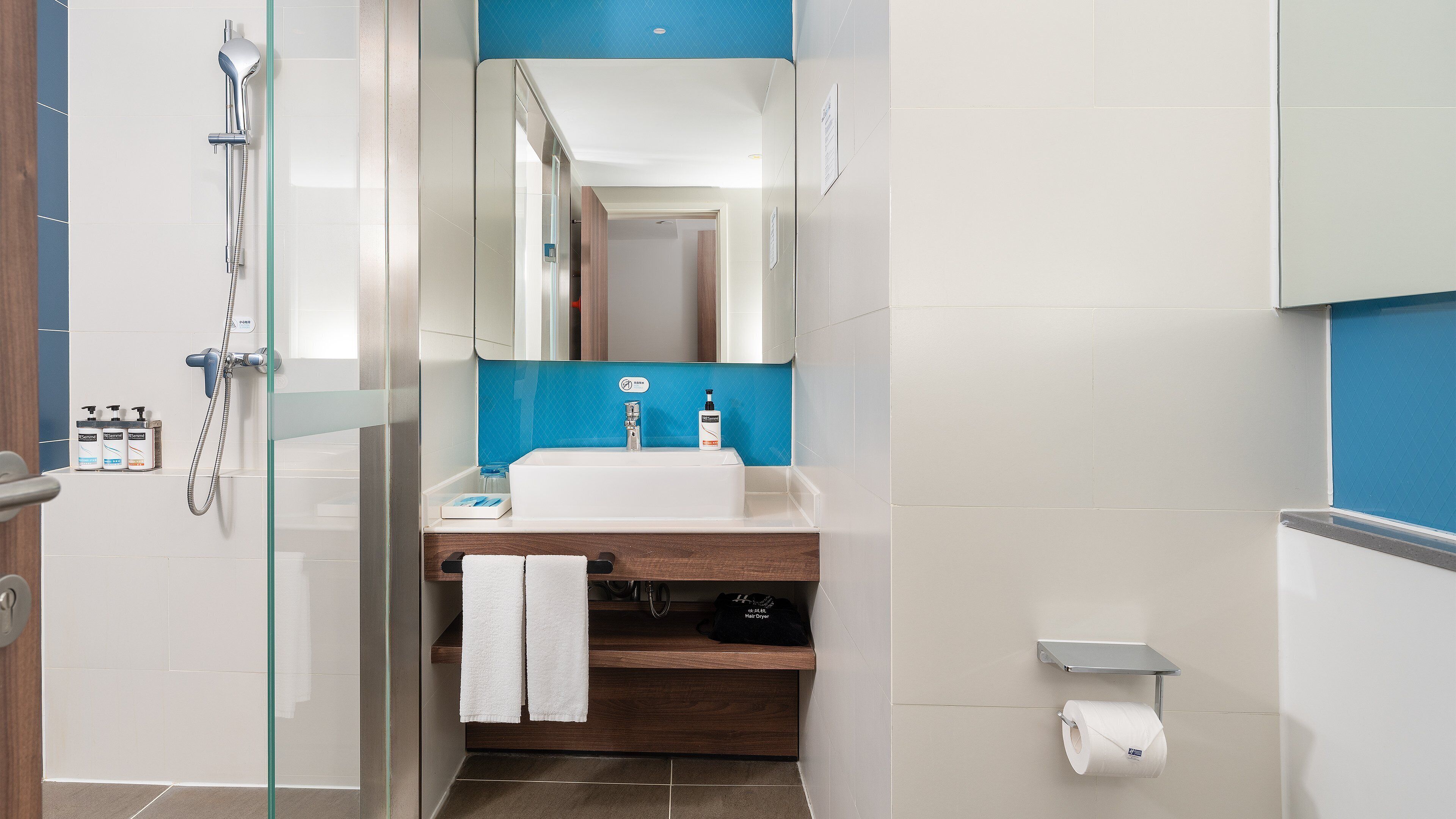 Un baño moderno con cabina de ducha de vidrio, un lavamanos blanco y una pared azul de acento, y un tocador de madera.