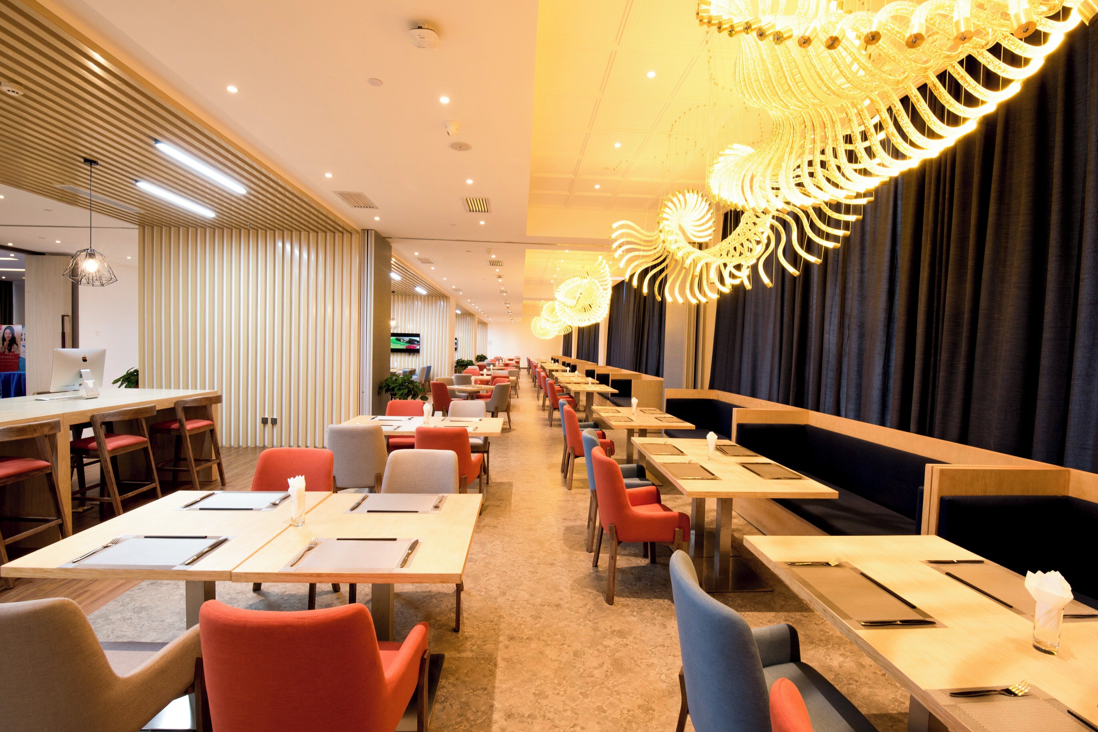 A hotel dining area with multiple tables and chairs, a television screen, and a decorative ceiling light fixture.