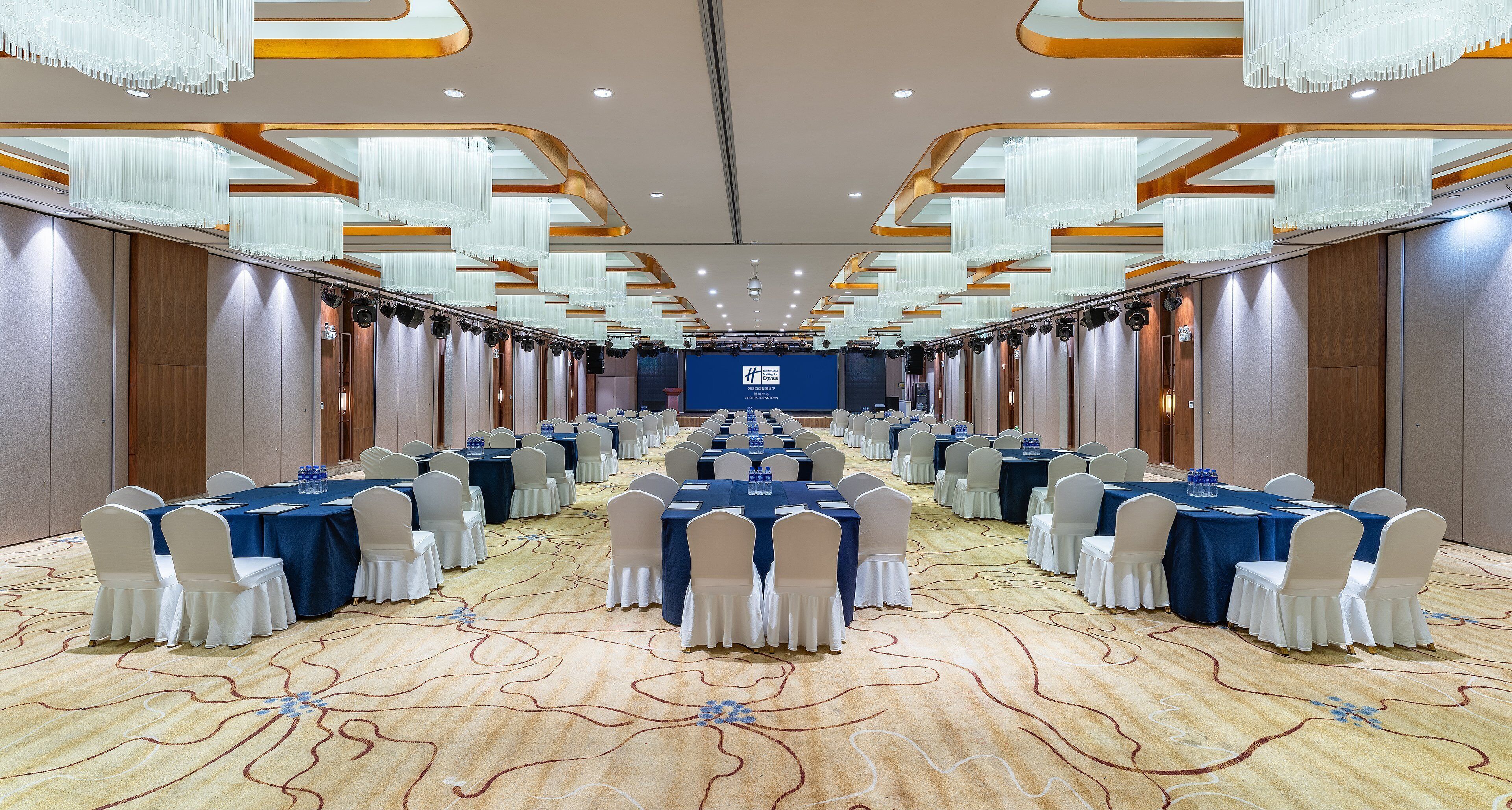 A conference room with rows of tables and chairs, a patterned carpet, and modern chandeliers.