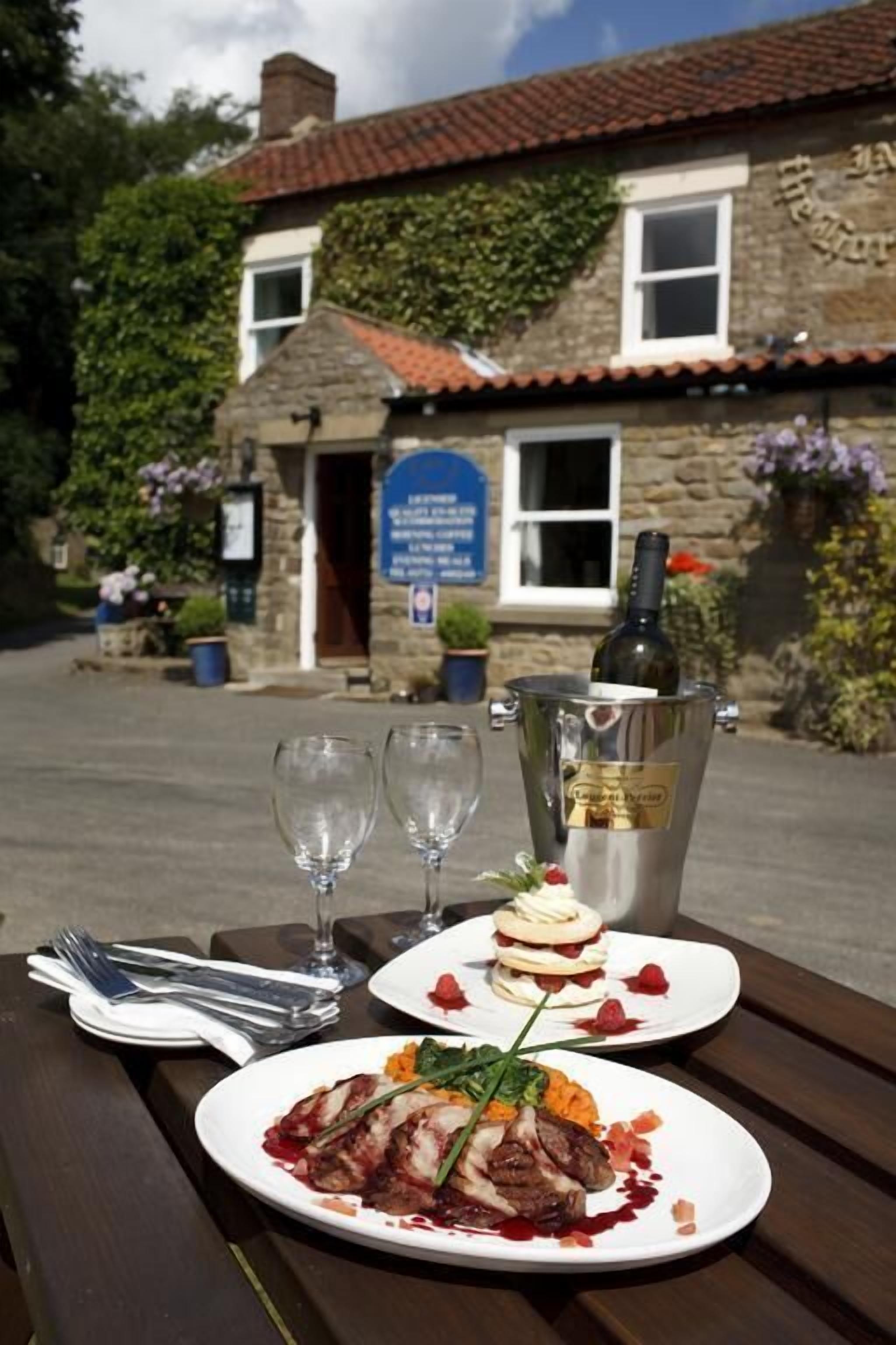 A dining table with plates of food, wine glasses, and a bottle of wine outside a stone building with a red-tiled roof.