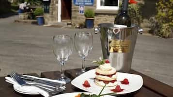 A dining table with plates of food, wine glasses, and a bottle of wine outside a stone building with a red-tiled roof.