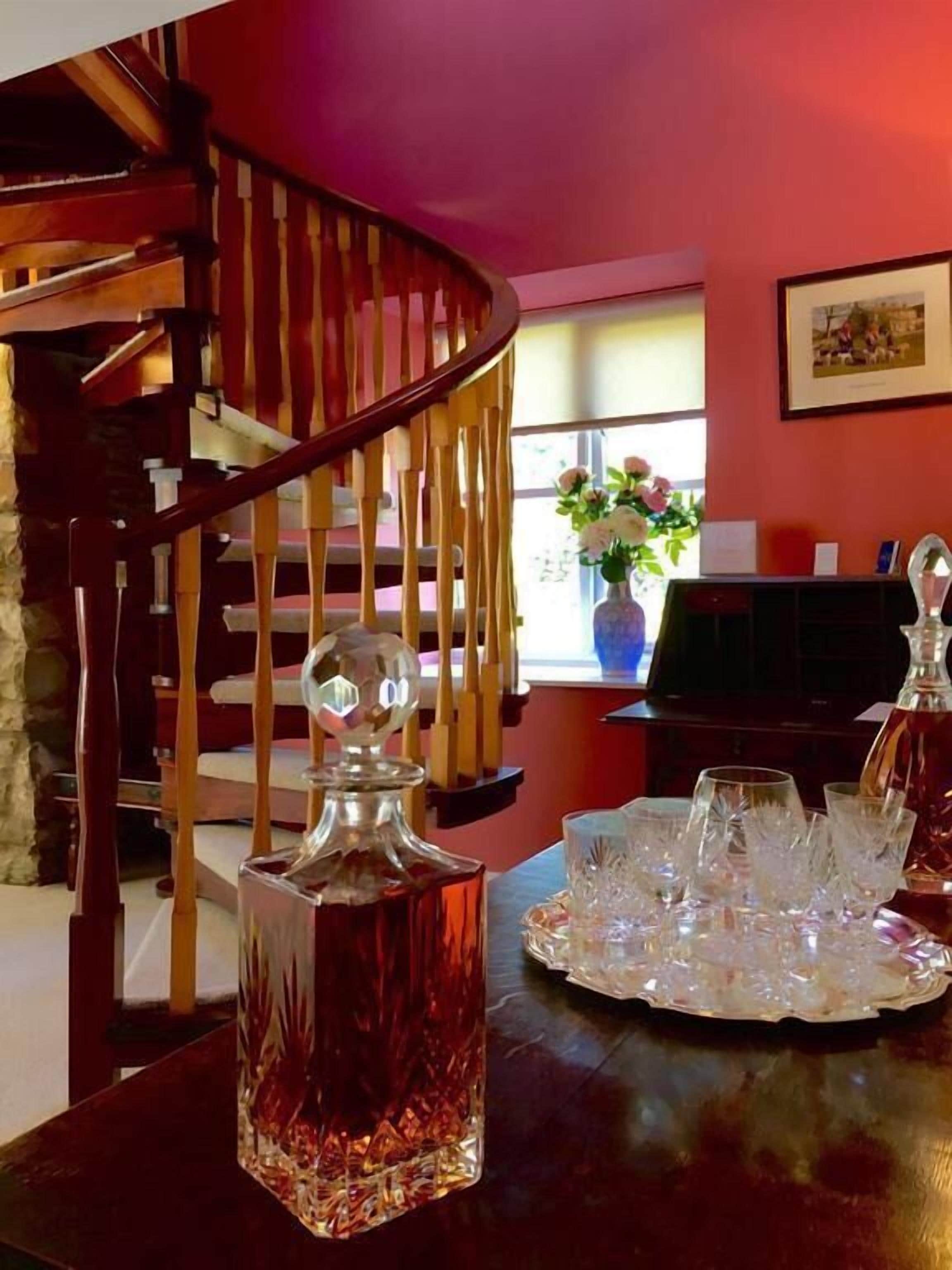 Una habitación con escalera de madera, chimenea y una mesa de comedor con un decantador de cristal y vasos.