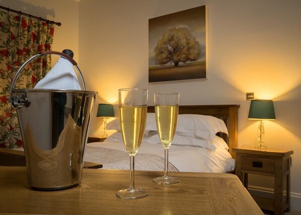 A hotel room with a bed, two bedside lamps, a wooden nightstand, a bucket with a cloth, and two glasses of champagne. - The Horseshoe Inn (Pickering)