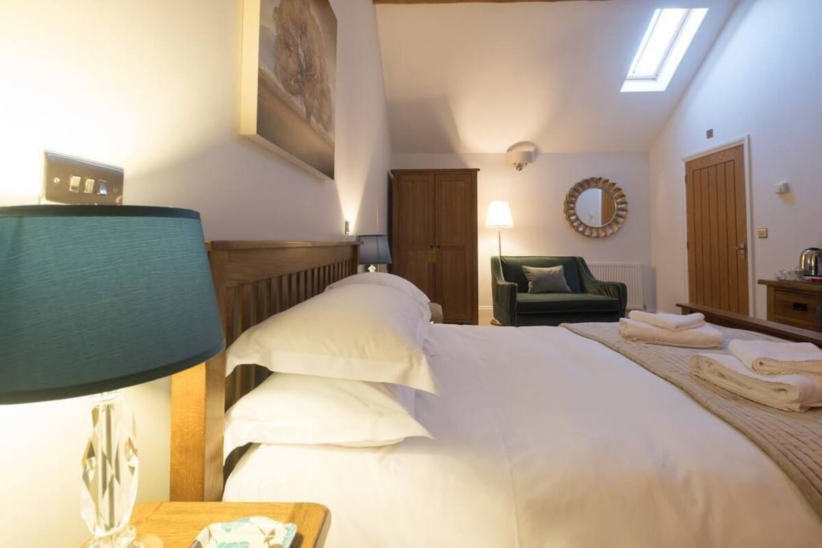 A hotel room with a large bed, a desk with a lamp, a chair, a wardrobe, and a skylight.