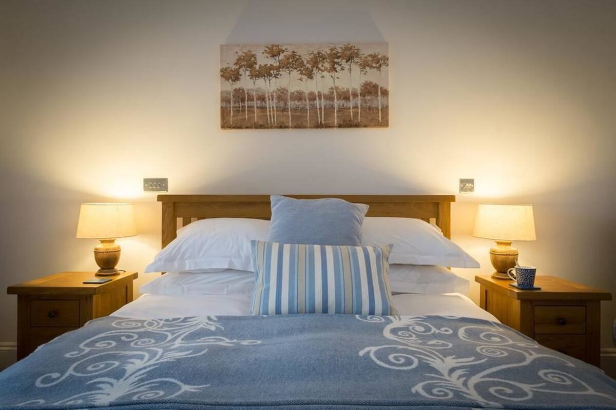 A neatly made bed with a blue patterned blanket, two bedside lamps, and a painting of trees on the wall.