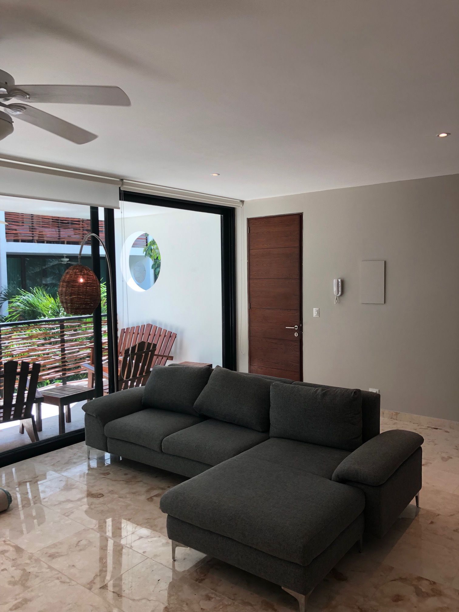 A modern living room with a grey sectional sofa, a ceiling fan, and a glass door leading to an outdoor area with a table and chairs.