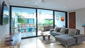 Ein modernes Wohnzimmer mit grauem Sofa, einem Holztisch und einem Esstisch mit blauen Stühlen. Eine Glastür öffnet sich zu einem Außenbereich mit einer Terrasse und Sitzgelegenheiten.