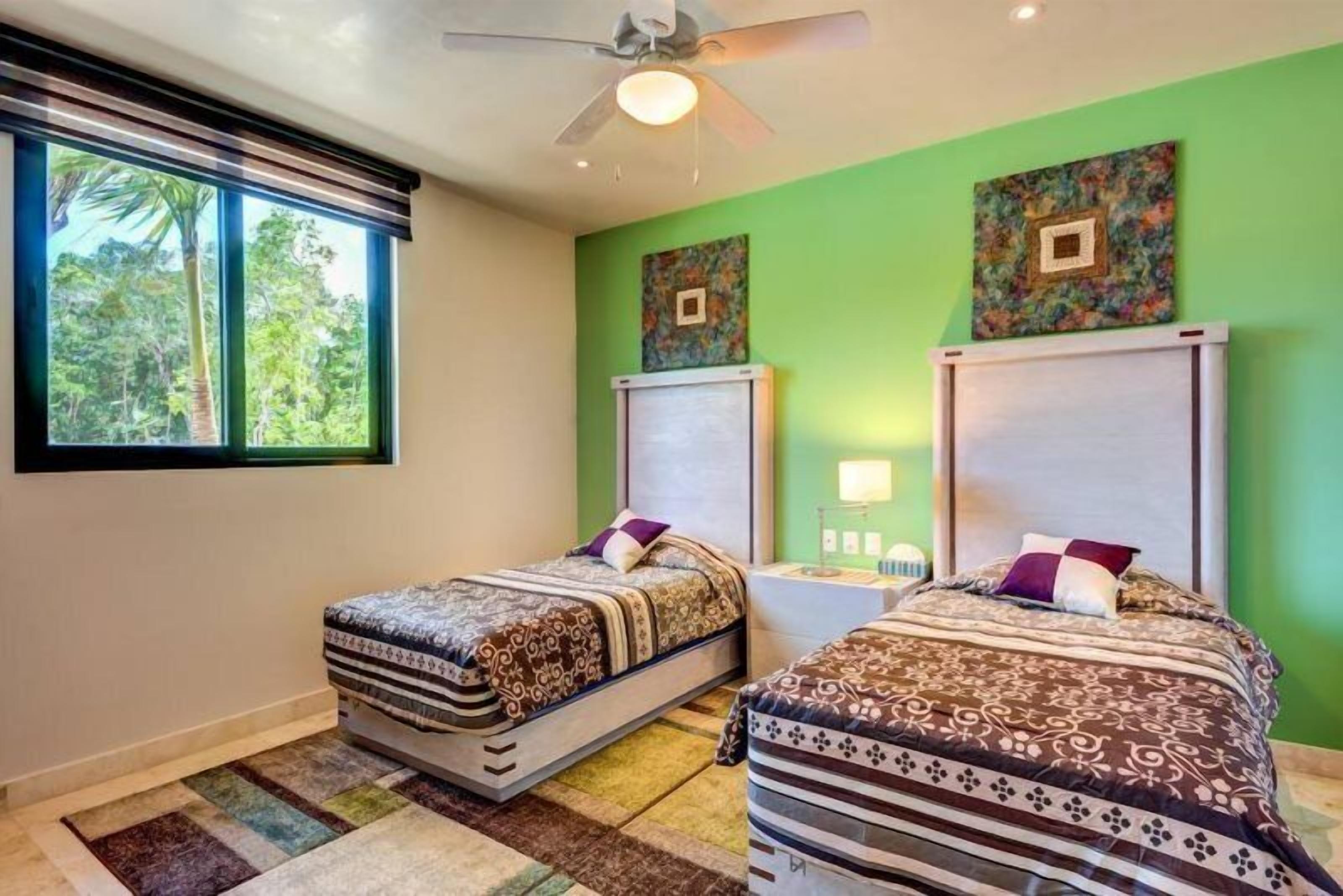 A room with two beds, a ceiling fan, a window with a view of trees, and artwork on the walls.