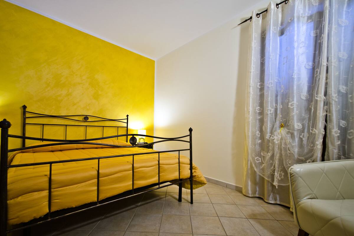 A room with a bed, a chair, a curtain, and a wall with a yellow accent.