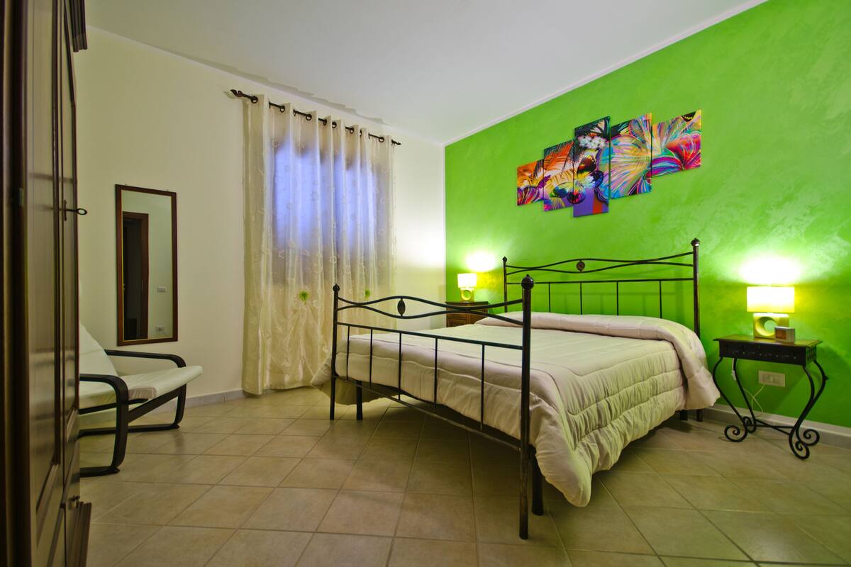 A bedroom with a bed, a chair, a nightstand, and a wall with a colorful painting.