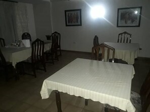 Un comedor con una gran mesa cubierta por un mantel blanco, rodeada de sillas de madera. Hay dos cuadros enmarcados en la pared sobre la mesa.