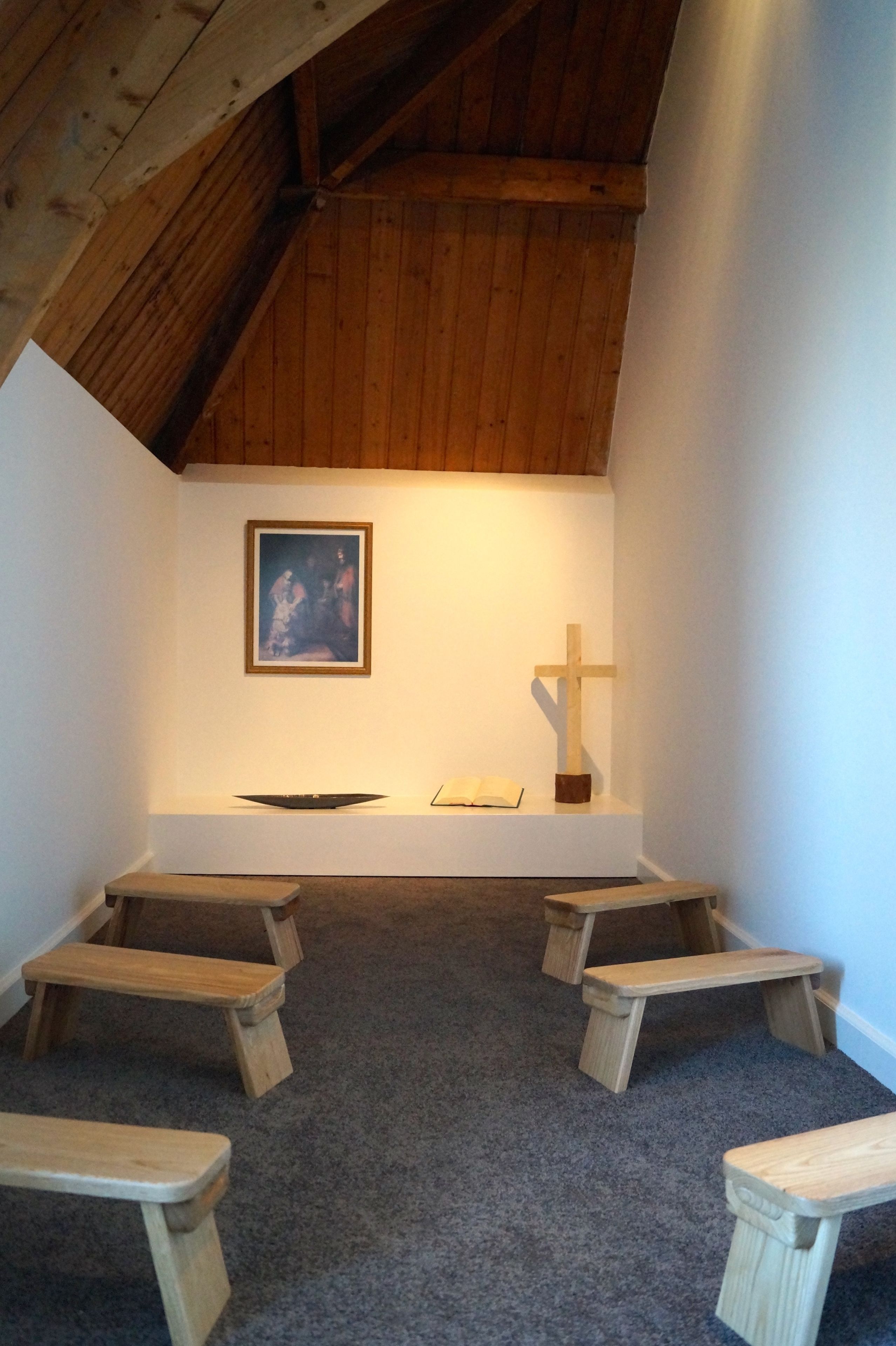 Une petite chapelle avec des bancs en bois, un crucifix en bois et un tableau encadré accroché au mur.