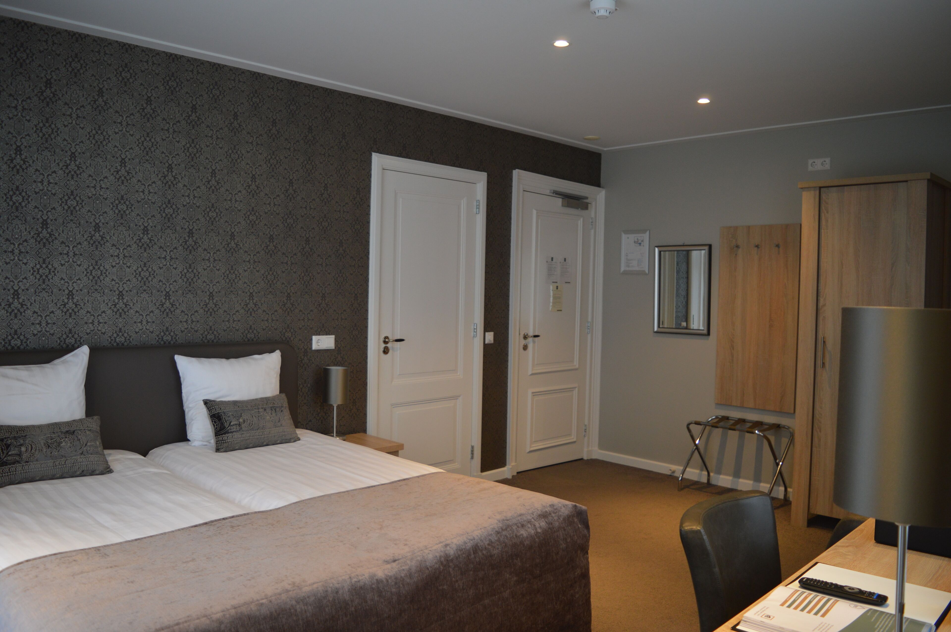 A hotel room with a large bed, two white doors, a wooden wardrobe, a desk with a lamp, and a chair.