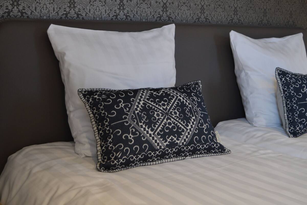 Een netjes opgemaakt bed met witte en gemêleerde kussens, een donker hoofdbord en een lamp op een nachtkastje.