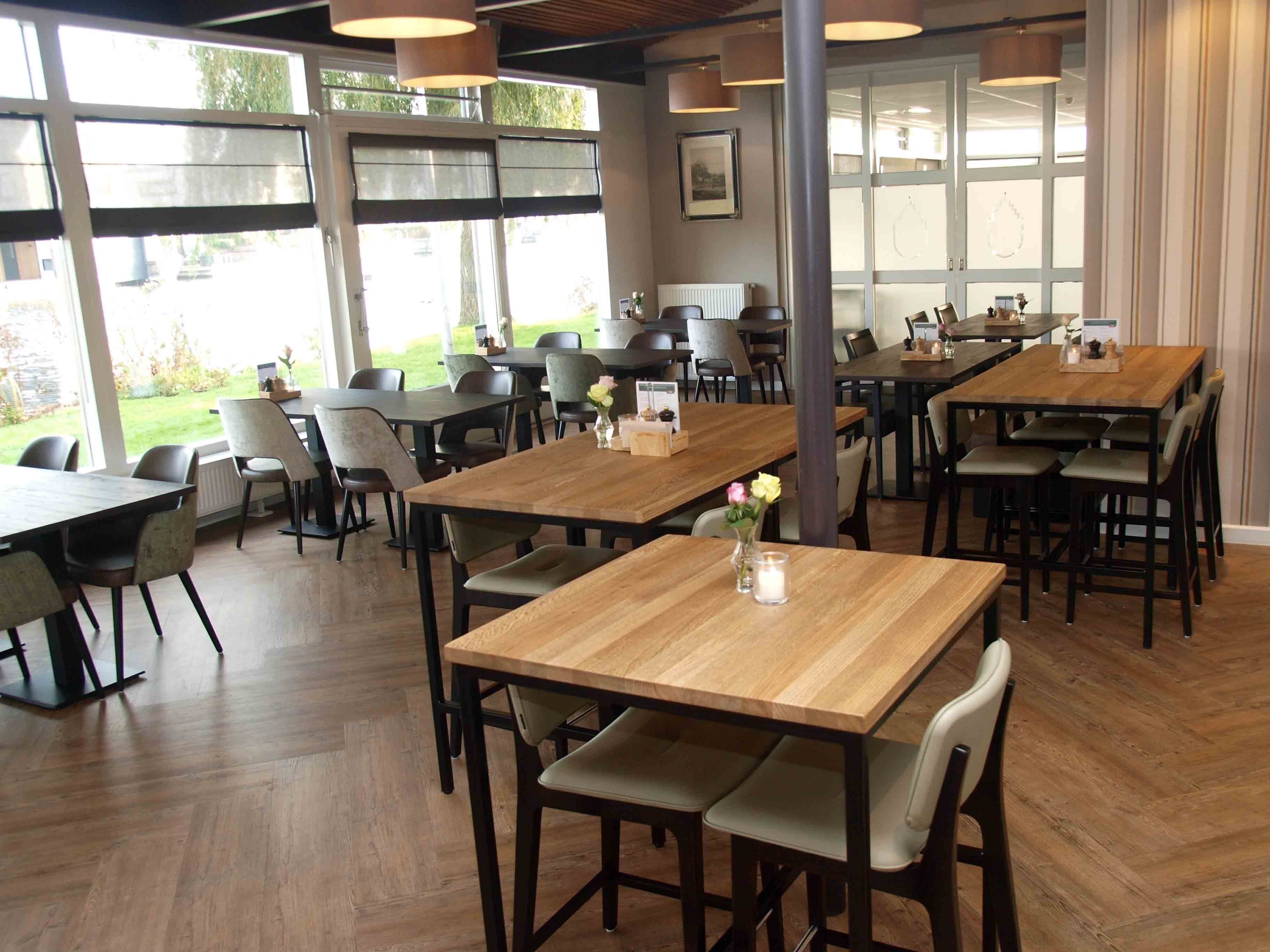 Un'ampia sala da pranzo con diversi tavoli e sedie in legno, ampie finestre che offrono vista sull'esterno e illuminazione a sospensione.