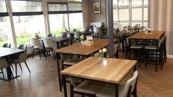 Un'ampia sala da pranzo con diversi tavoli e sedie in legno, ampie finestre che offrono vista sull'esterno e illuminazione a sospensione.