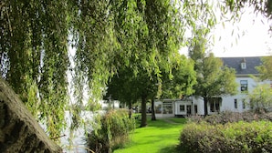 Een rustige rivierentuin met een wit gebouw, een stenen muur en weelderige begroeiing.