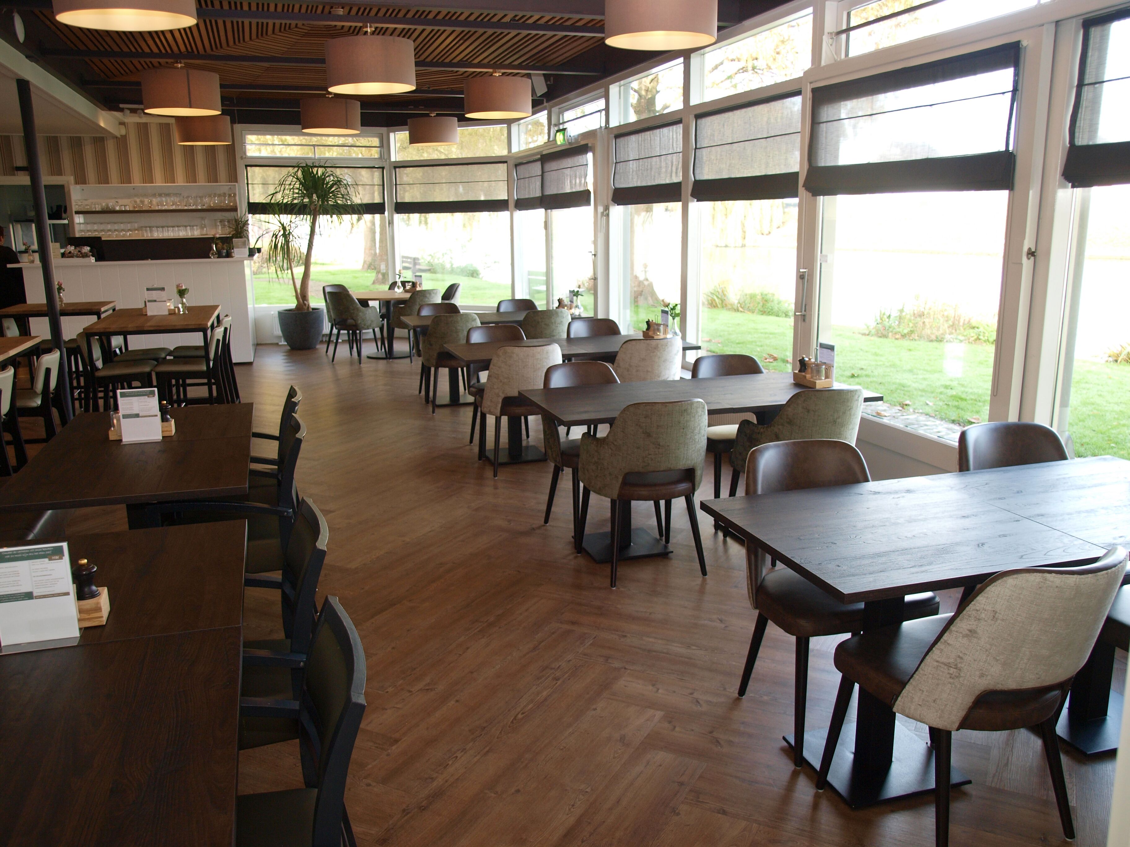 Un restaurante con mesas y sillas de madera, ventanas amplias y lámparas colgantes.