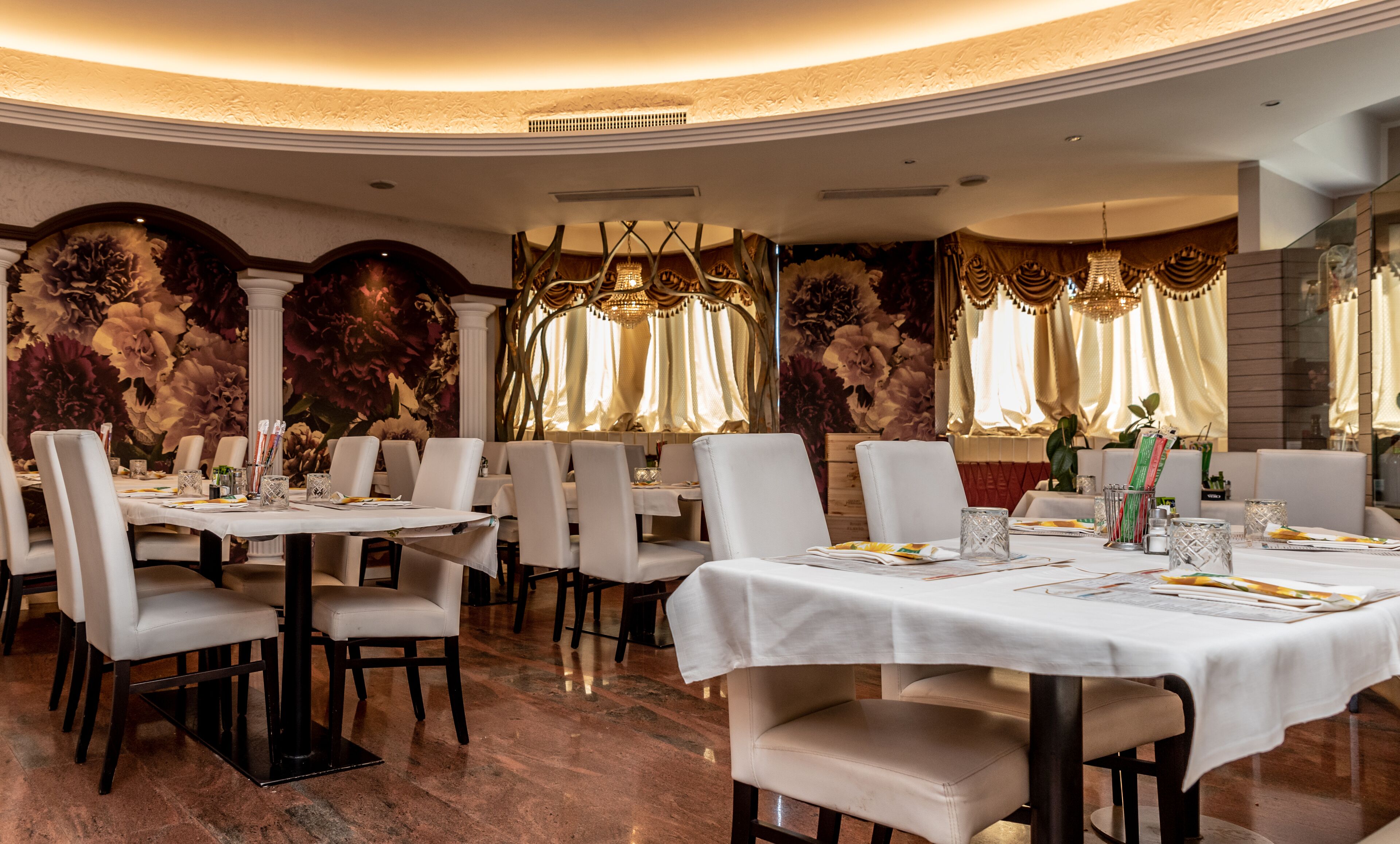 A restaurant with round tables, white tablecloths, and a floral mural on the walls.