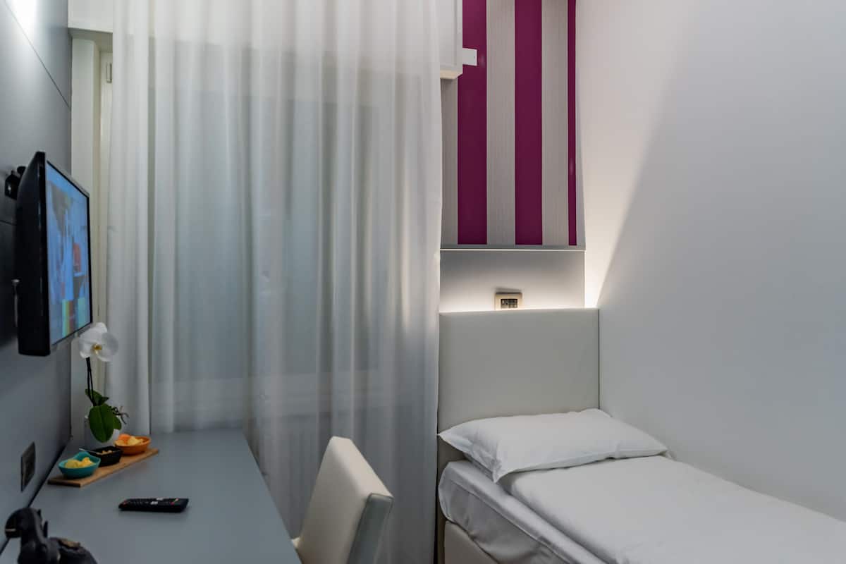 Une chambre d’hôtel avec un lit, un bureau et une chaise, une télévision, une petite table avec un vase de fleurs et une fenêtre avec des rideaux.