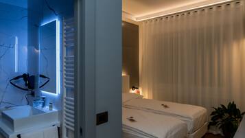 Habitación de hotel con baño que incluye un lavamanos moderno y un espejo, cama con ropa blanca y una mesita de noche con lámpara.